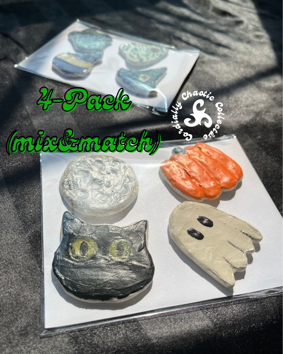 A 4-pack of handmade Halloween-themed fridge magnets displayed on a dark textured fabric background. The set includes a black cat head, an orange pumpkin, a beige ghost, and a textured silver moon, together in their own clear plastic sleeve.