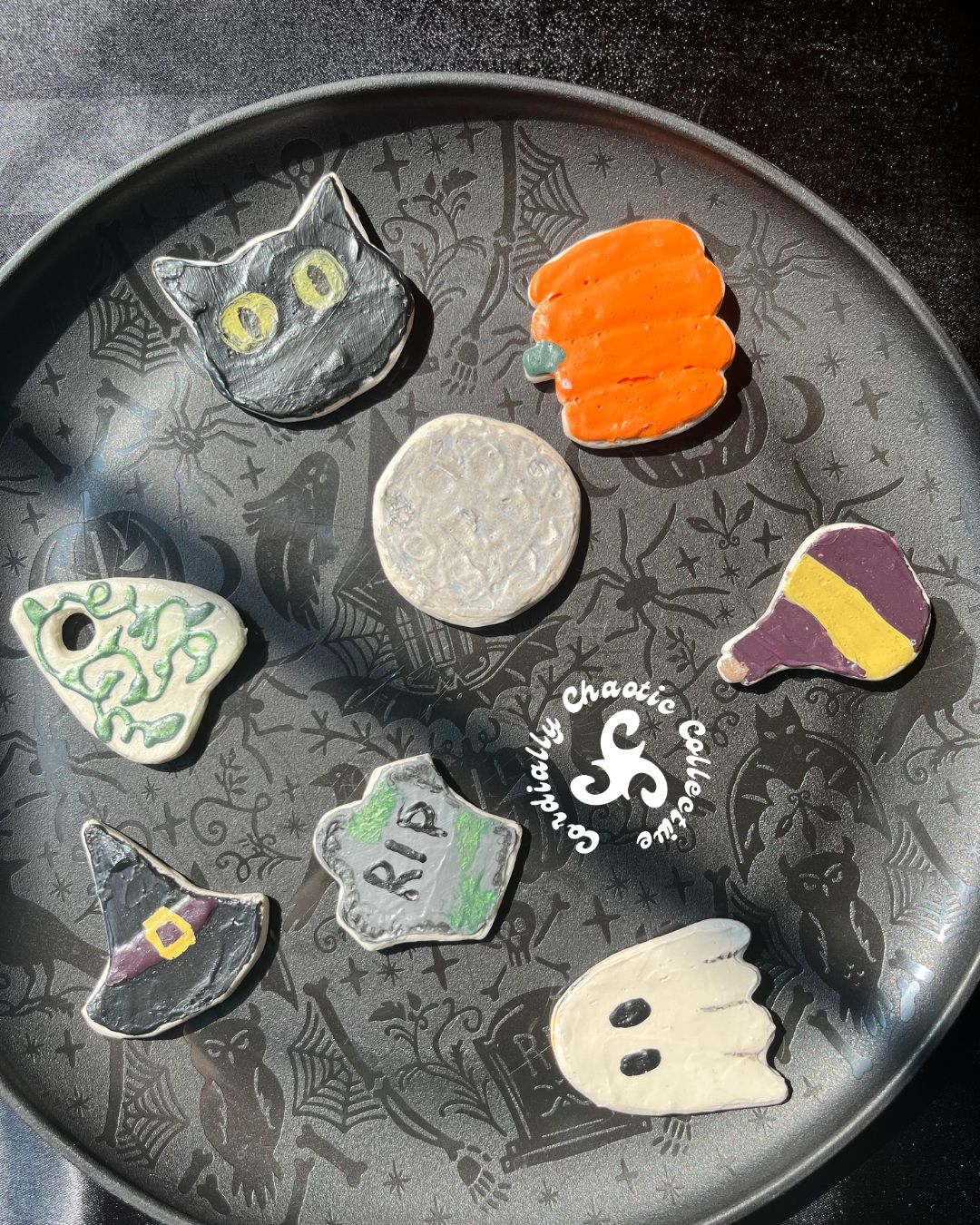 A collection of seven hand-painted ceramic Halloween magnets arranged on a dark patterned tray. The charms include a black cat with yellow eyes, an orange pumpkin, a silver full moon, a green ivy planchette, a purple potion bottle, a black witch hat with a gold buckle, a gray tombstone reading 'RIP', and a white ghost.